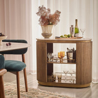 A modern bar cart with a fluted wood finish and open shelves, styled in a bright dining room with a vase of flowers, wine, and glasses on top.