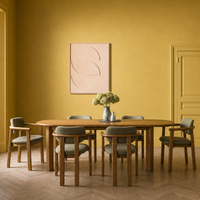 A dining room scene with a round wooden table and six upholstered chairs in a warm mustard-yellow room. A framed abstract print hangs on the wall above the table, and a vase with flowers sits at the center.