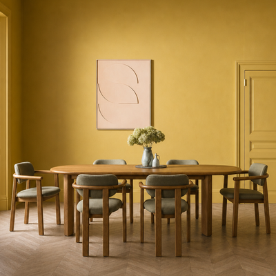 A dining room scene with a round wooden table and six upholstered chairs in a warm mustard-yellow room. A framed abstract print hangs on the wall above the table, and a vase with flowers sits at the center.