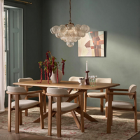 A dining room scene with a light wood dining table and six upholstered chairs, styled with a glass bubble chandelier, a vase of red flowers, and a framed abstract painting on a muted green wall.