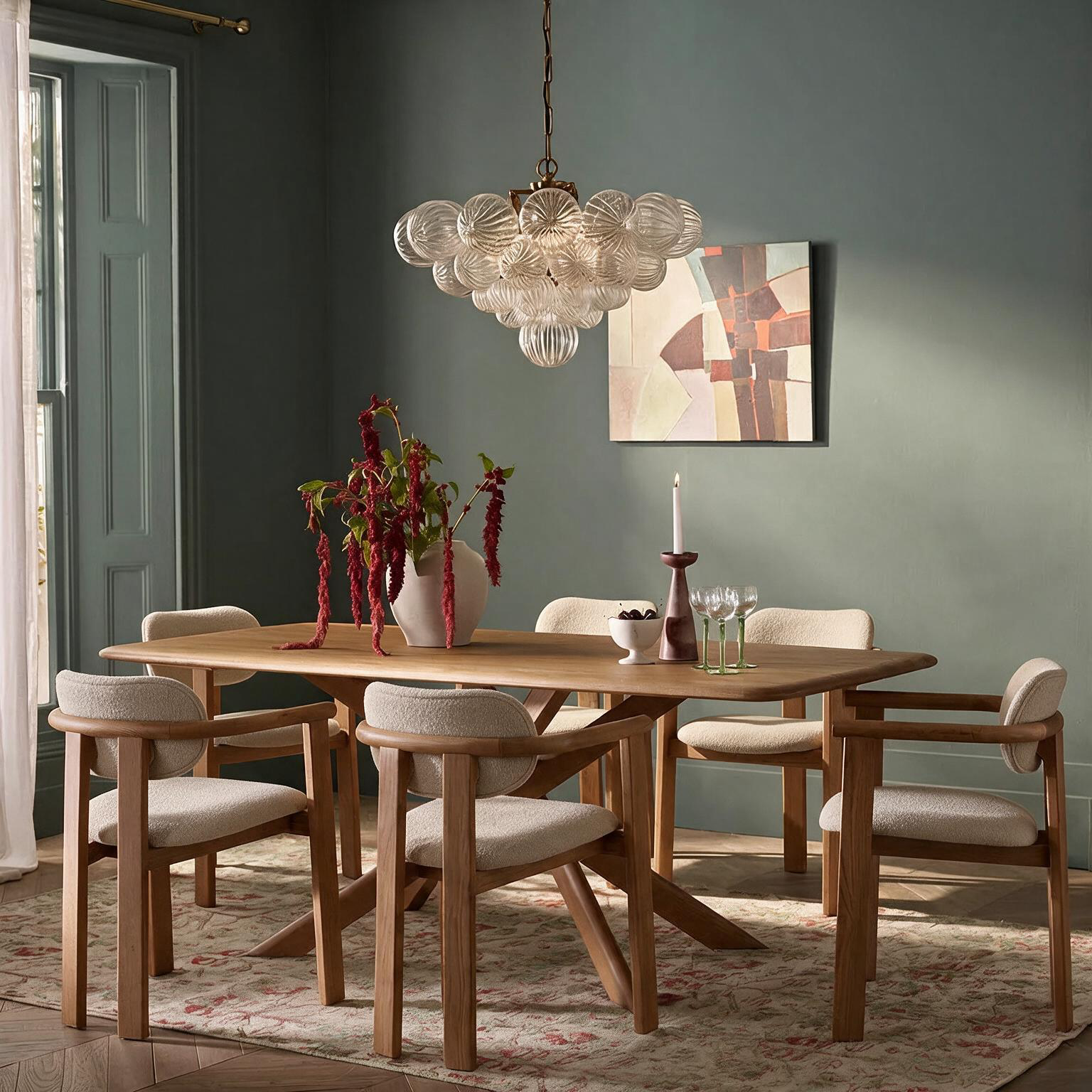 A dining room scene with a light wood dining table and six upholstered chairs, styled with a glass bubble chandelier, a vase of red flowers, and a framed abstract painting on a muted green wall.