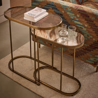 Two nested oval nesting side tables with gold metal frames and glass tops, styled in a living room setting with a patterned sofa and a rug.