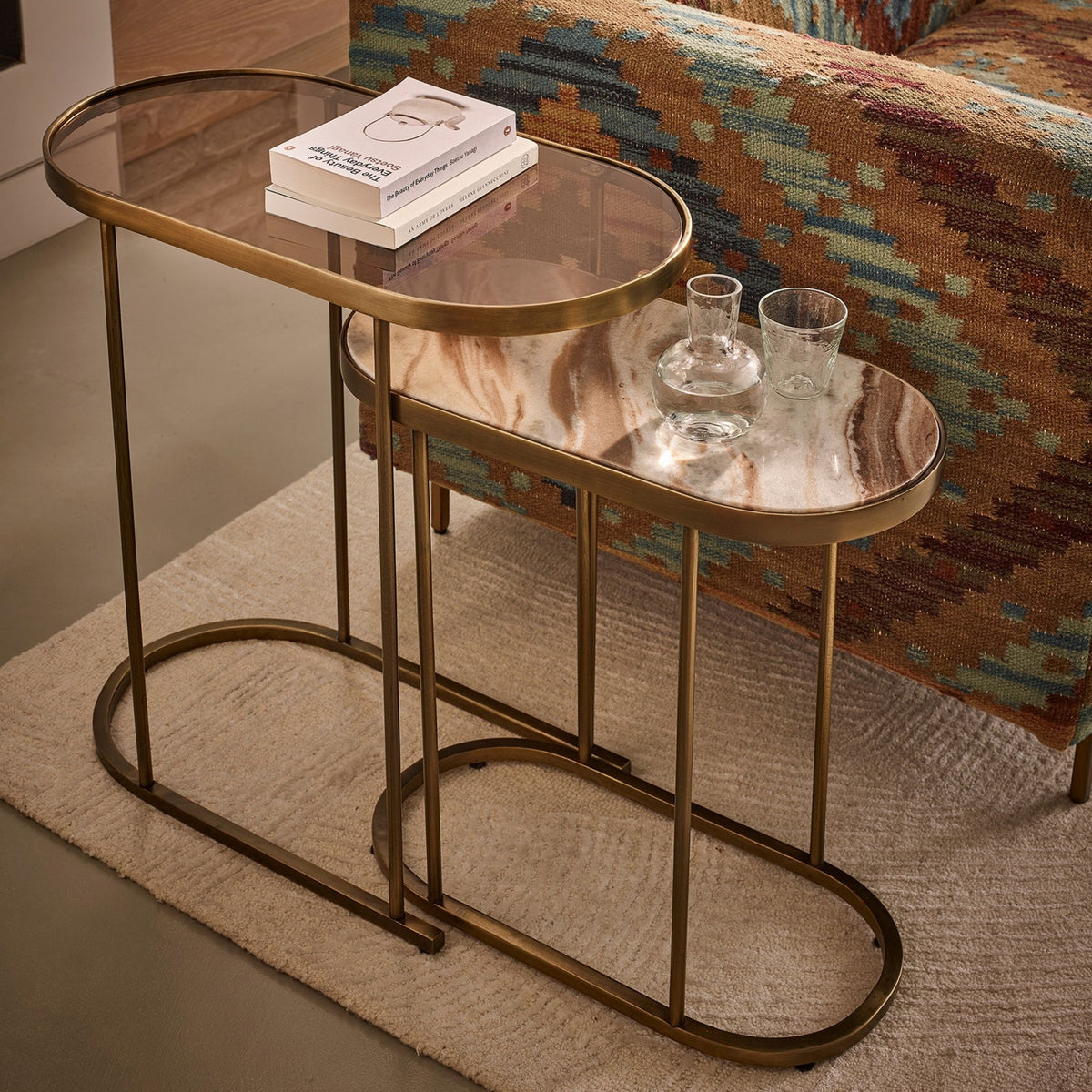 Two nested oval nesting side tables with gold metal frames and glass tops, styled in a living room setting with a patterned sofa and a rug.