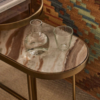 A small oval side table with a gold metal frame and legs, featuring a polished beige and brown marble-patterned top. Two clear glass drinking vessels sit on the tabletop: a rounded decanter and a short tumbler.