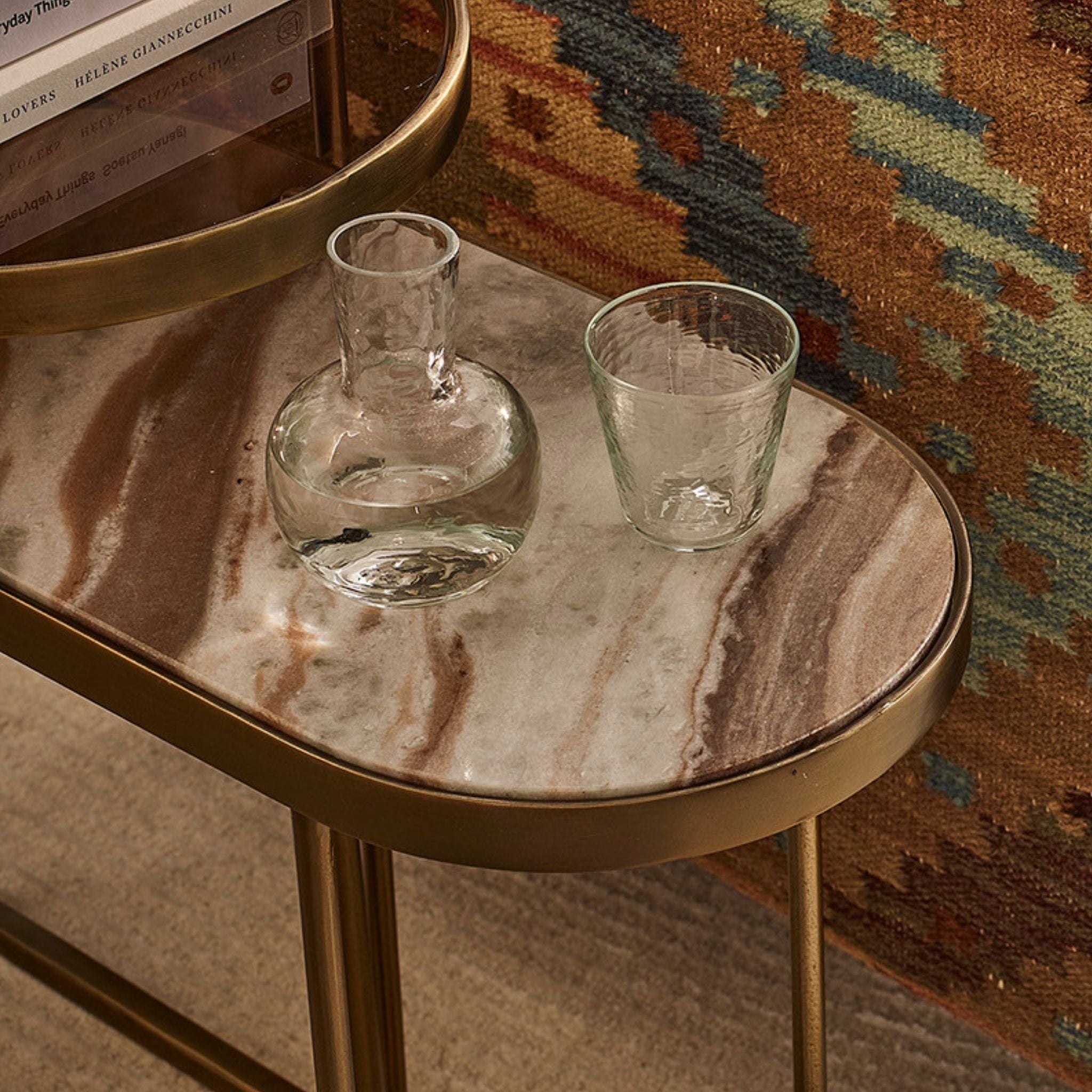 A small oval side table with a gold metal frame and legs, featuring a polished beige and brown marble-patterned top. Two clear glass drinking vessels sit on the tabletop: a rounded decanter and a short tumbler.