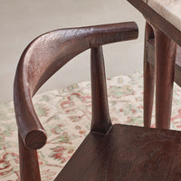 Close-up of a dark brown wooden dining chair with a curved armrest and textured grain, positioned over a patterned rug next to another wooden table.