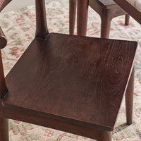 Close-up of a dark brown wooden chair seat with visible wood grain, set on a patterned rug with other wooden furniture partially visible in the background.