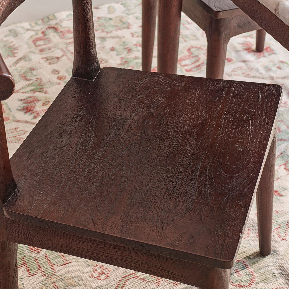 Close-up of a dark brown wooden chair seat with visible wood grain, set on a patterned rug with other wooden furniture partially visible in the background.
