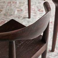 Close-up of a dark brown wooden chair with a curved backrest and textured grain, set on a patterned rug with other furniture legs visible in the background.