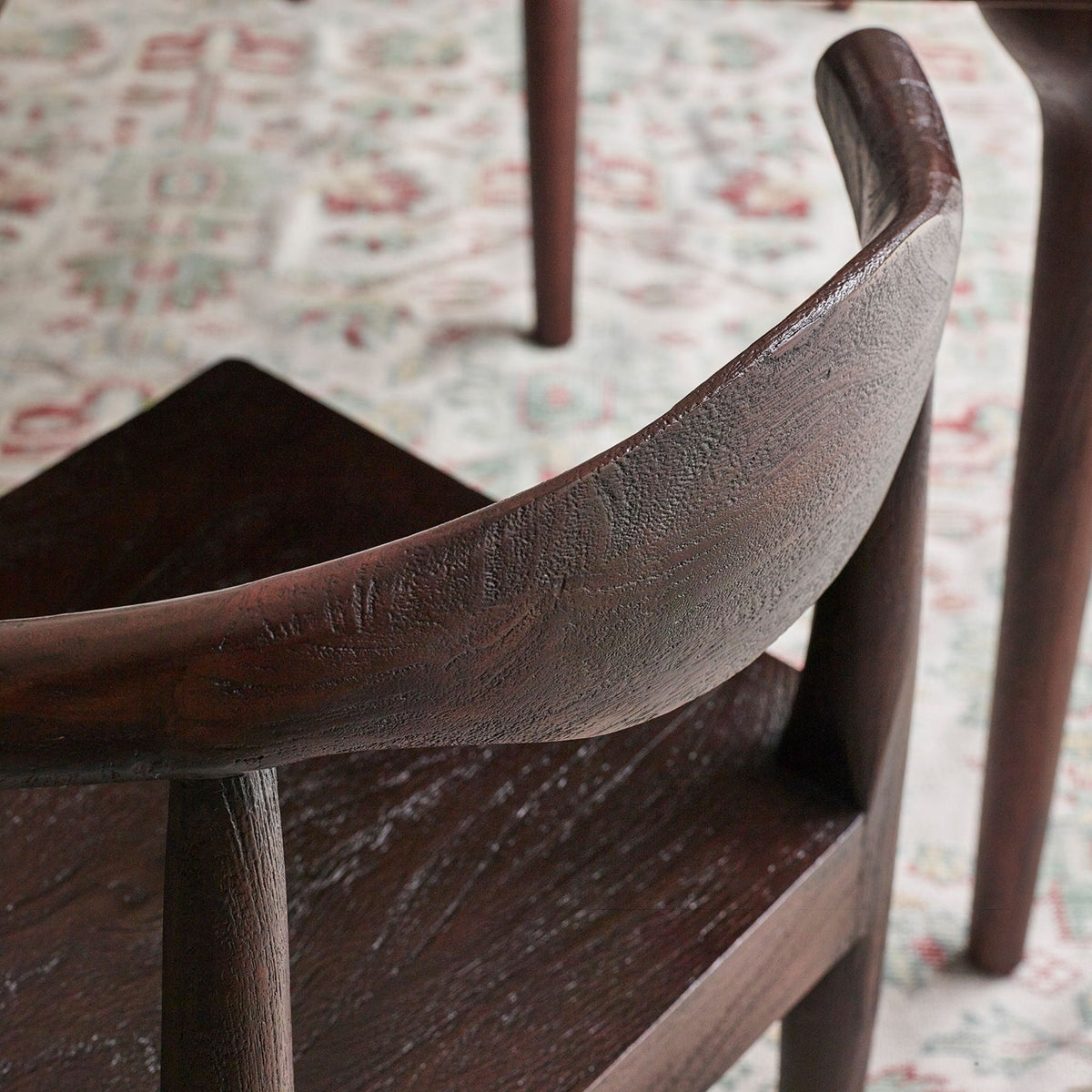 Close-up of a dark brown wooden chair with a curved backrest and textured grain, set on a patterned rug with other furniture legs visible in the background.