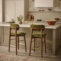 Two mid-century modern bar stools with olive green velvet seats and wooden frames, positioned at a kitchen island in a warm, contemporary kitchen setting.