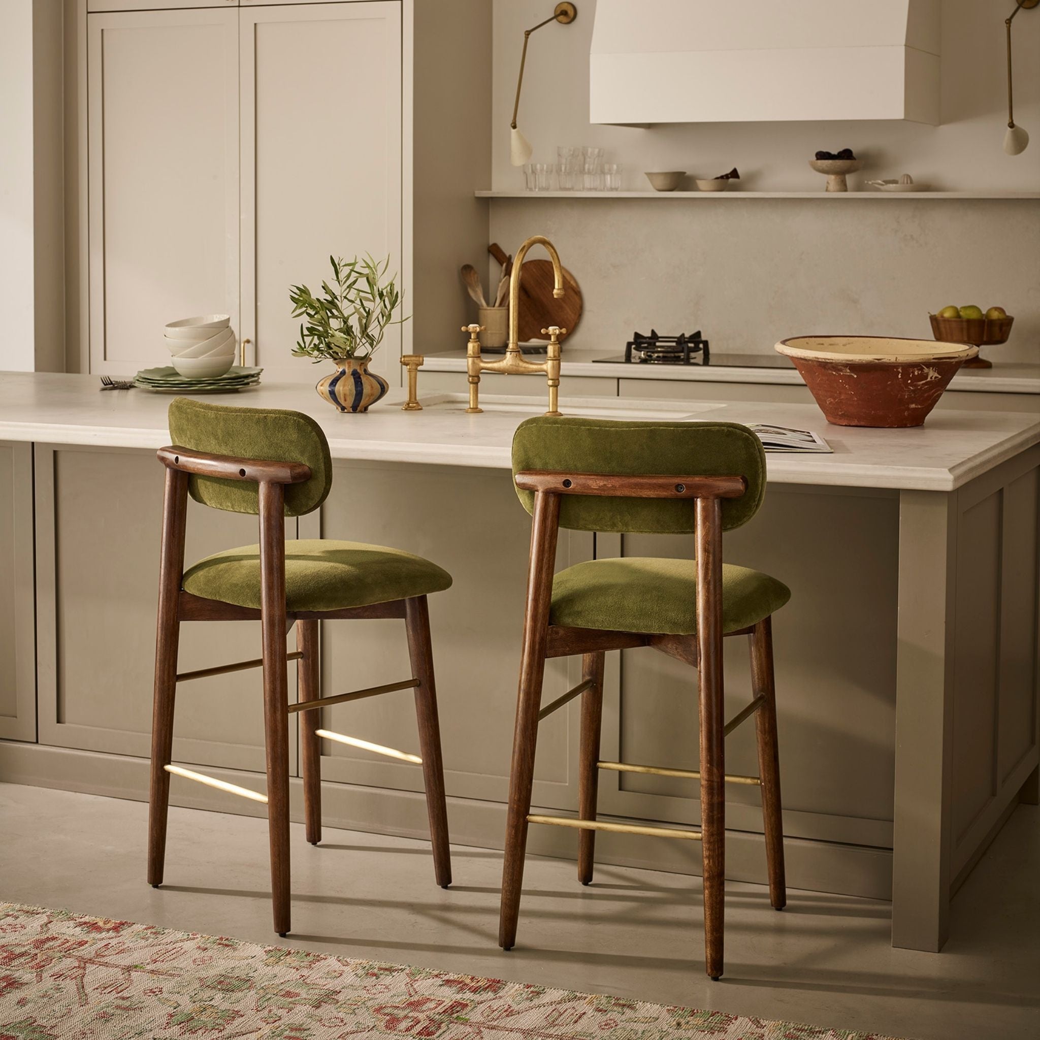 Two mid-century modern bar stools with olive green velvet seats and wooden frames, positioned at a kitchen island in a warm, contemporary kitchen setting.