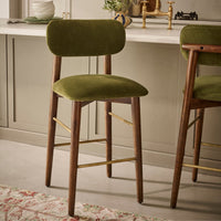 An olive green velvet bar stool with a wooden frame and brass-colored metal accents, set in a modern kitchen environment.