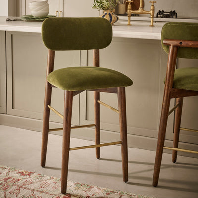 An olive green velvet bar stool with a wooden frame and brass-colored metal accents, set in a modern kitchen environment.