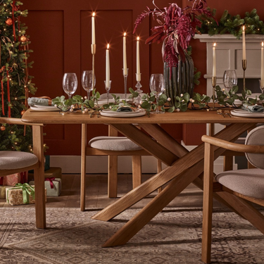 A wooden dining table with a unique crisscross X base and matching upholstered chairs, styled for a holiday dinner in a warm, decorated room with a Christmas tree, fireplace, and garlands.