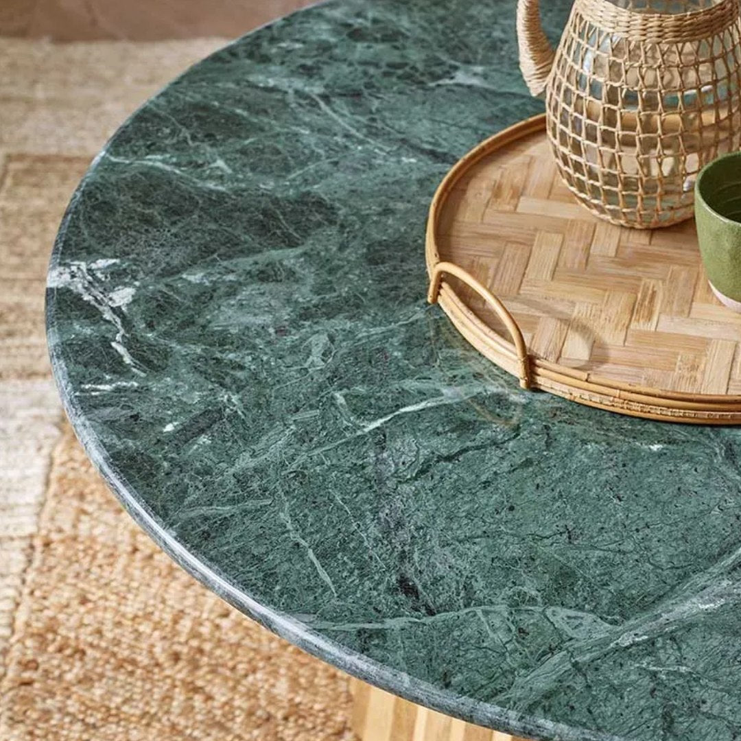 A round coffee table with a dark green marble top and a fluted light wood base, styled with a woven tray and wicker pitcher on a beige rug.